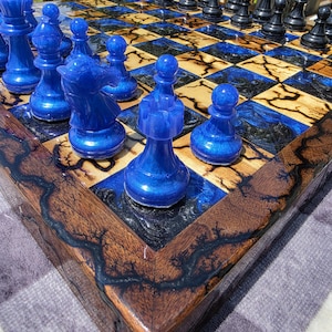 May include: A chess set featuring a wooden board with dark, lightning-like fractal patterns and blue resin squares. The pieces consist of solid black figures on one side and translucent, shimmering blue figures on the other, including pawns, rooks, and knights.