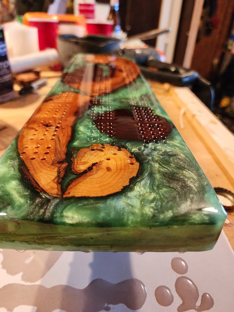 May include: A handcrafted cribbage board with a green epoxy resin base, featuring inlaid wood pieces. The board has drilled holes for pegs and a smooth, glossy finish. The wood pieces are a light brown color.