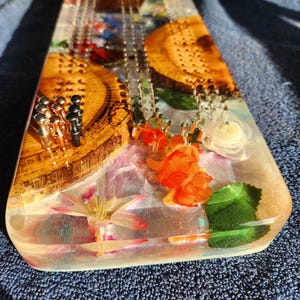 May include: A handcrafted cribbage board made of wood and resin. The board features embedded flowers, wood slices, and metal pegs. The resin is clear, allowing the decorative elements to be visible. The board is rectangular with rounded corners.
