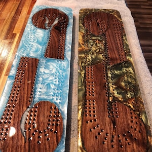 May include: Two wooden cribbage boards with inlaid resin. One board has a blue and white resin, the other has a gold and green resin. Both boards have a dark wood playing surface with metal peg holes.