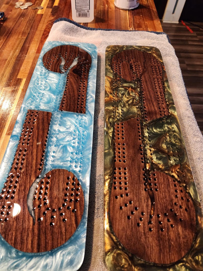 May include: Two wooden cribbage boards with inlaid resin. One board has a blue and white resin, the other has a gold and green resin. Both boards have a dark wood playing surface with metal peg holes.