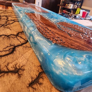 May include: A rectangular wooden serving board with a blue resin river design. The wood has a natural grain, and the resin has a swirling, ocean-like appearance. The board is approximately 30 inches long.