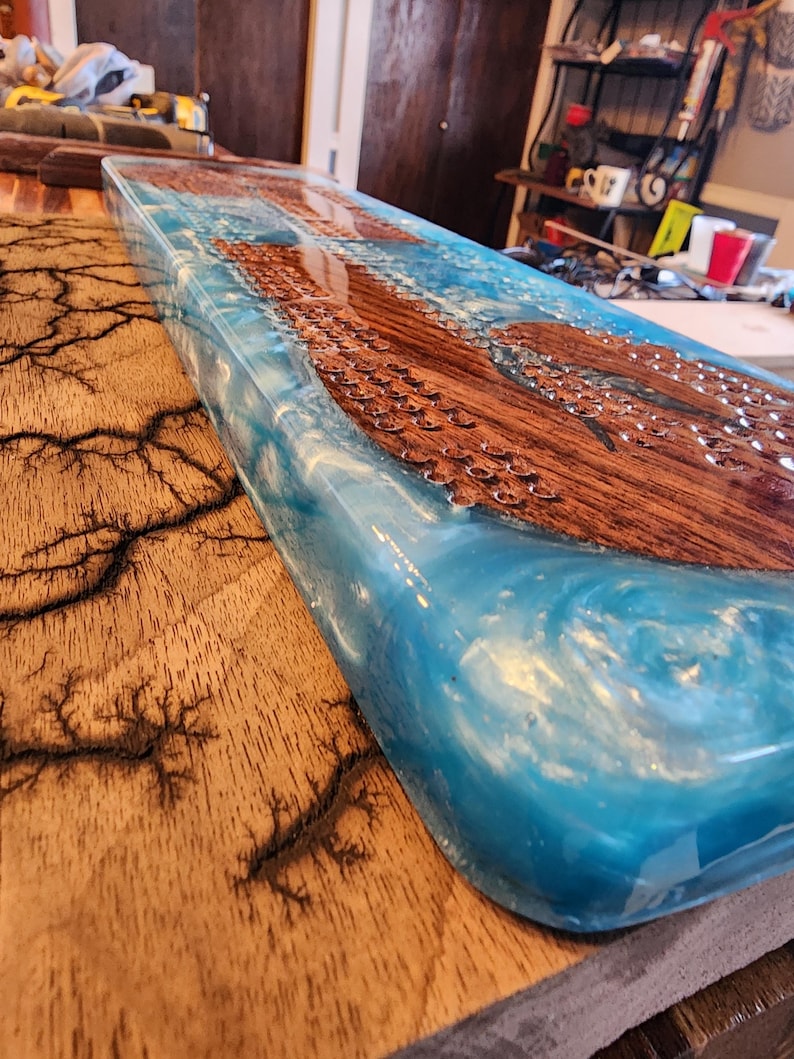 May include: A rectangular wooden serving board with a blue resin river design. The wood has a natural grain, and the resin has a swirling, ocean-like appearance. The board is approximately 30 inches long.