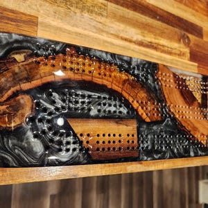 May include: A handcrafted cribbage board made of wood and black and white resin. The board features a unique design with a wooden inlay and drilled holes for pegs. The wood has a natural grain and the resin has a marbled effect.