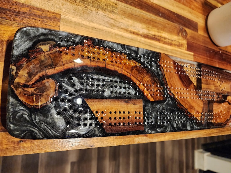 May include: A handcrafted cribbage board made of wood and black and white resin. The board features a unique design with a wooden inlay and drilled holes for pegs. The wood has a natural grain and the resin has a marbled effect.