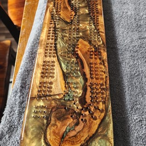 May include: A handcrafted cribbage board made of wood and epoxy resin. The board features a natural wood grain pattern with areas of gold and green resin. Numerous small holes are drilled for peg placement, creating a unique and functional game board.
