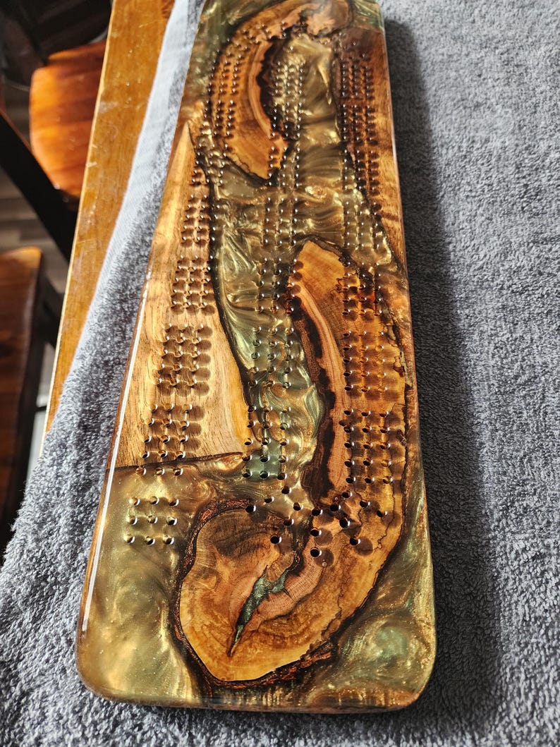 May include: A handcrafted cribbage board made of wood and epoxy resin. The board features a natural wood grain pattern with areas of gold and green resin. Numerous small holes are drilled for peg placement, creating a unique and functional game board.