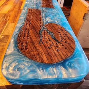 May include: A handcrafted cribbage board with a wooden playing surface and a vibrant blue resin border. The board features drilled holes for pegs and a unique design, perfect for game nights. The board measures approximately 24 inches long.