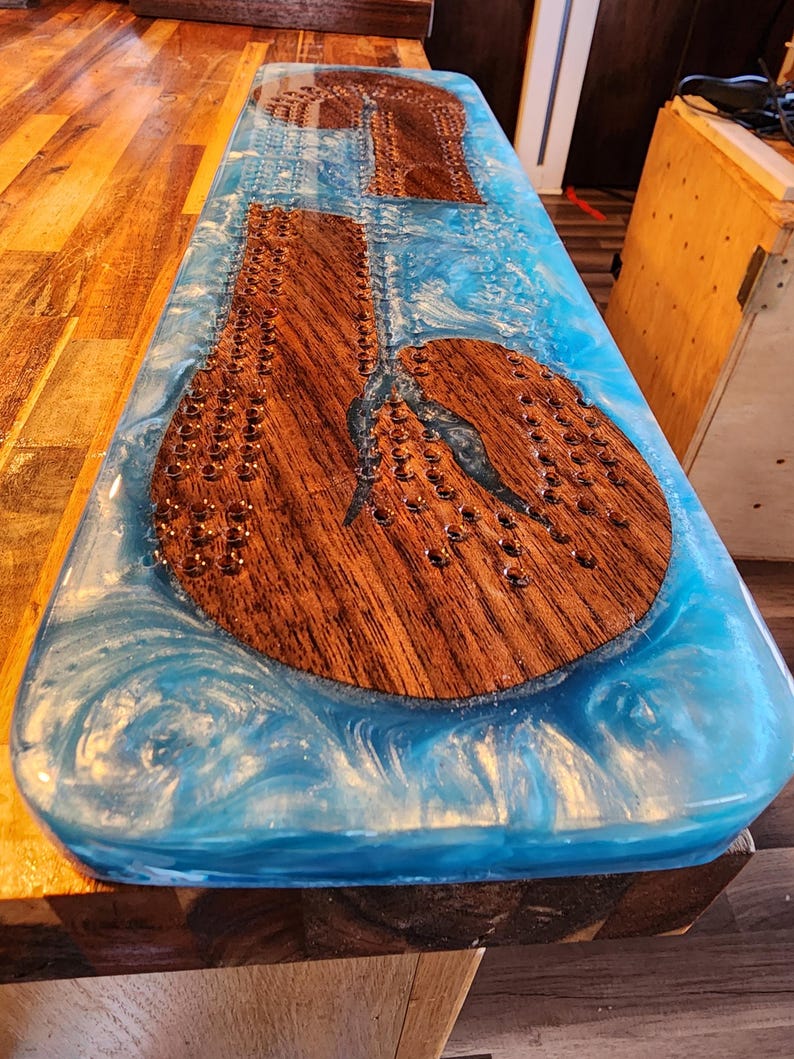 May include: A handcrafted cribbage board with a wooden playing surface and a vibrant blue resin border. The board features drilled holes for pegs and a unique design, perfect for game nights. The board measures approximately 24 inches long.