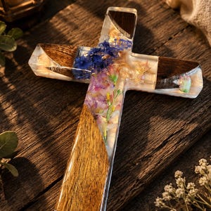May include: A handcrafted wooden cross with a clear resin center, inlaid with dried flowers in shades of purple, blue, and white. The cross arms and base are made of dark wood, creating a striking contrast. The resin has a glossy finish.