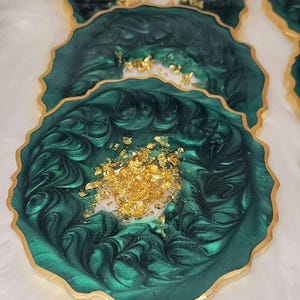 May include: Emerald green resin coasters with gold leaf accents and gold trim. The coasters have a unique, organic shape and are arranged on a white, fluffy surface. The text "Emerald Green" is visible at the top.