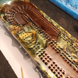 May include: A handcrafted cribbage board with a wooden playing surface and embedded metal pegs. The board is encased in a clear resin with swirls of gold, green, and brown. A blue resin block is visible in the background.
