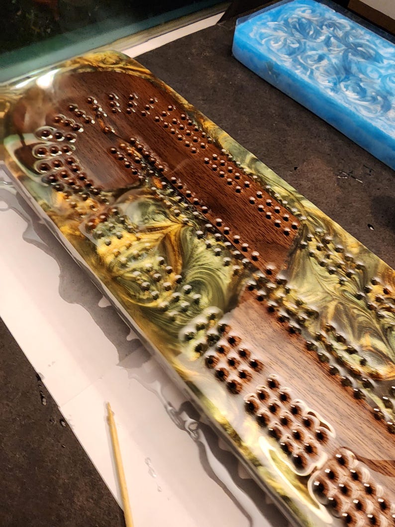 May include: A handcrafted cribbage board with a wooden playing surface and embedded metal pegs. The board is encased in a clear resin with swirls of gold, green, and brown. A blue resin block is visible in the background.