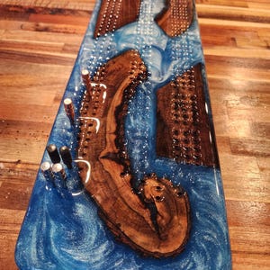 May include: A rectangular cribbage board with a blue epoxy resin river design. The board features inlaid wooden pieces and metal pegs. The wood has a natural grain, and the resin has a swirling, ocean-like appearance.