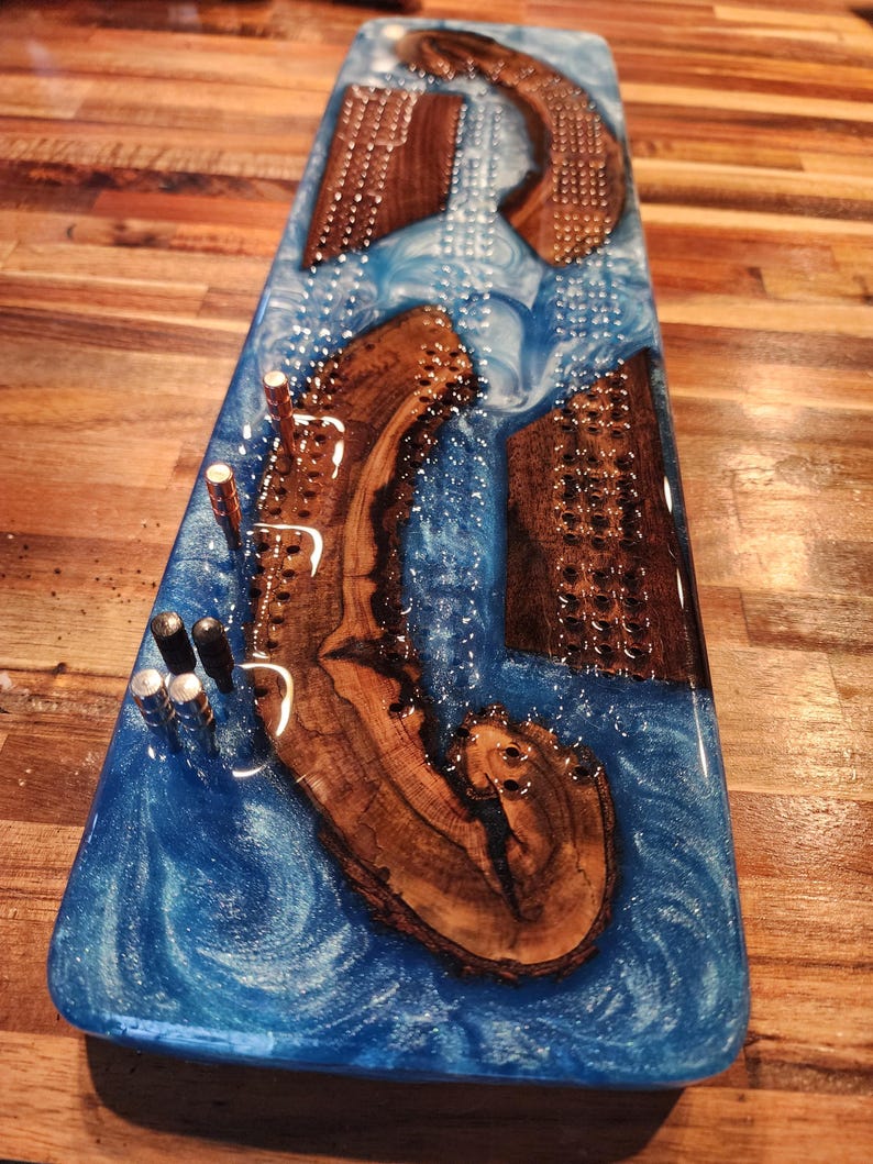 May include: A rectangular cribbage board with a blue epoxy resin river design. The board features inlaid wooden pieces and metal pegs. The wood has a natural grain, and the resin has a swirling, ocean-like appearance.