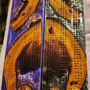 May include: Two wooden cribbage boards with a unique design. One board has a purple resin base, the other a gray resin base. Both boards feature a natural wood grain pattern and drilled holes for pegs. A beautiful and functional game.