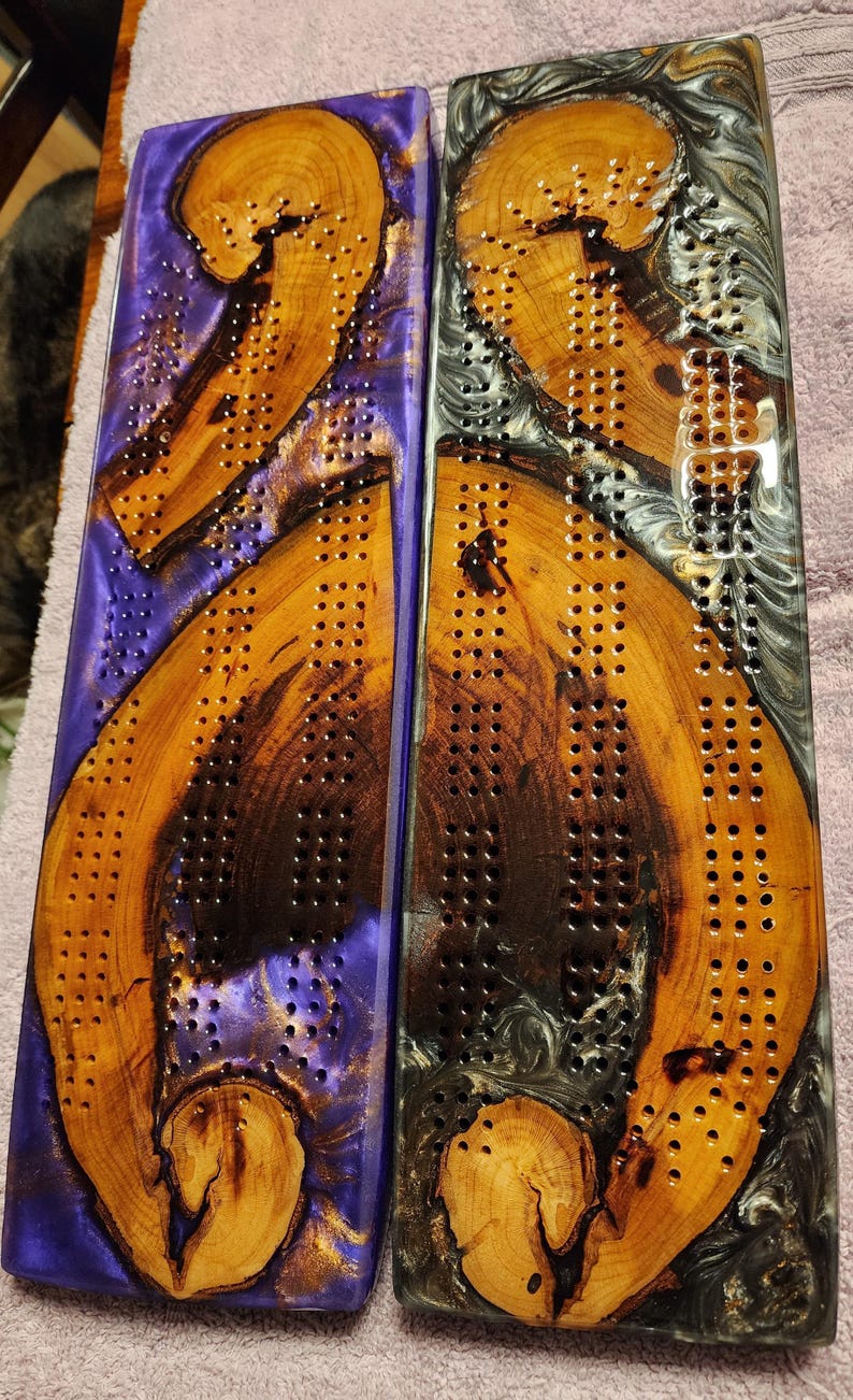 May include: Two wooden cribbage boards with a unique design. One board has a purple resin base, the other a gray resin base. Both boards feature a natural wood grain pattern and drilled holes for pegs. A beautiful and functional game.