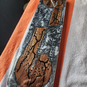 May include: A rectangular cribbage board with a wooden inlay design and a clear resin finish. The board features a dark wood grain pattern with black accents and small holes for pegs. The resin has a swirling gray and white pattern.