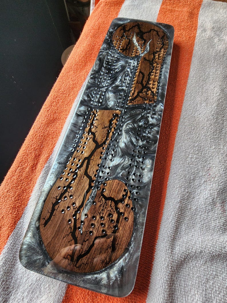 May include: A rectangular cribbage board with a wooden inlay design and a clear resin finish. The board features a dark wood grain pattern with black accents and small holes for pegs. The resin has a swirling gray and white pattern.