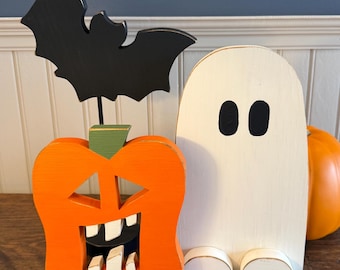 May include: A wooden orange jack-o'-lantern with a green stem and a black bat cutout attached to the top, positioned next to a white wooden ghost figure with black oval eyes and two small white feet, with a round orange pumpkin behind it.