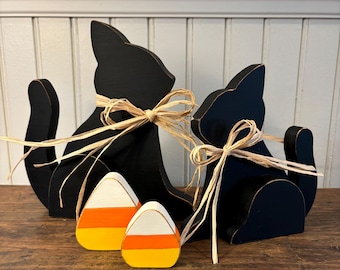 May include: Two black cat silhouettes with raffia bows, and two candy corn decorations. The wooden cats are in a sitting position. The candy corn decorations are white, orange, and yellow. Halloween-themed decor.