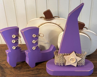 May include: Wooden Halloween decorations including purple witch boots and a hat, with a burlap and star accent. Two white pumpkins with twine stems are in the background. The items are on a wooden surface.