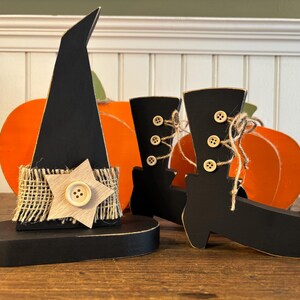 May include: Wooden Halloween decorations featuring a black witch hat with a star accent and a pair of black witch boots. The hat has a burlap band and a wooden star with a button. Orange pumpkin cutouts are in the background.