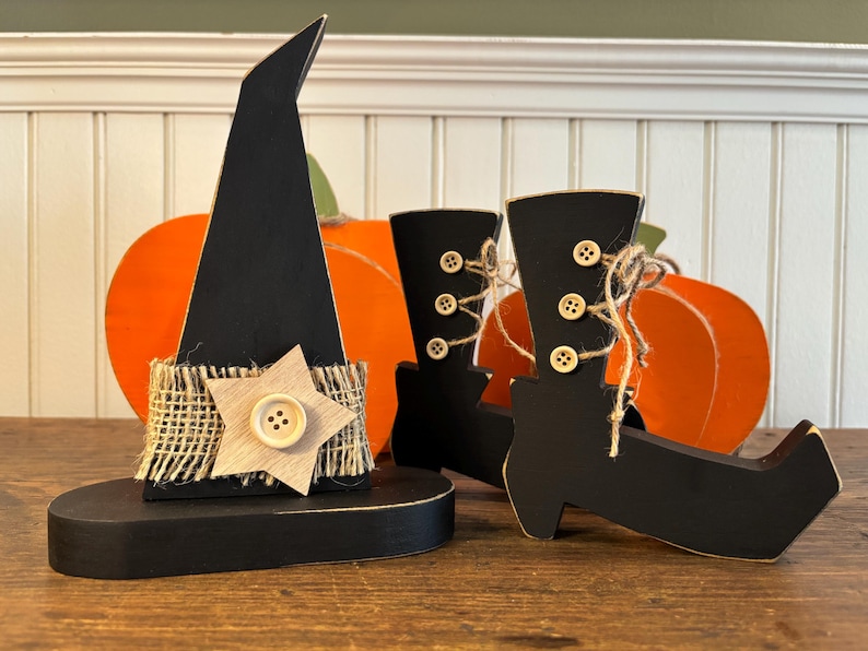 May include: Wooden Halloween decorations featuring a black witch hat with a star accent and a pair of black witch boots. The hat has a burlap band and a wooden star with a button. Orange pumpkin cutouts are in the background.