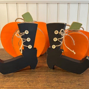 May include: Wooden Halloween decorations featuring black witch boots with button details and twine accents. Two orange pumpkin cutouts with green stems are in the background. The boots are a festive addition to fall decor.