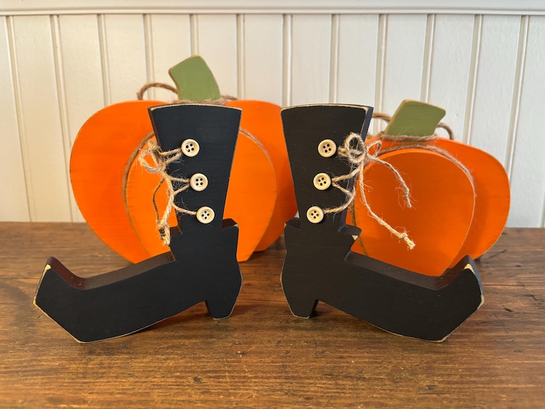 May include: Wooden Halloween decorations featuring black witch boots with button details and twine accents. Two orange pumpkin cutouts with green stems are in the background. The boots are a festive addition to fall decor.