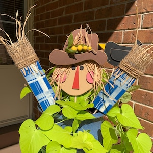 May include: A decorative scarecrow figure with a straw hat, button eyes, and a red nose. The scarecrow has blue plaid arms and is surrounded by bright green foliage in a blue pot. The background is a brick wall.