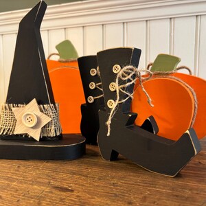 May include: Wooden Halloween decorations including a black witch hat with a star, black witch boots, and orange pumpkins. The hat has a burlap band with a wooden star and button. The boots have button details and twine. The pumpkins have green stems.