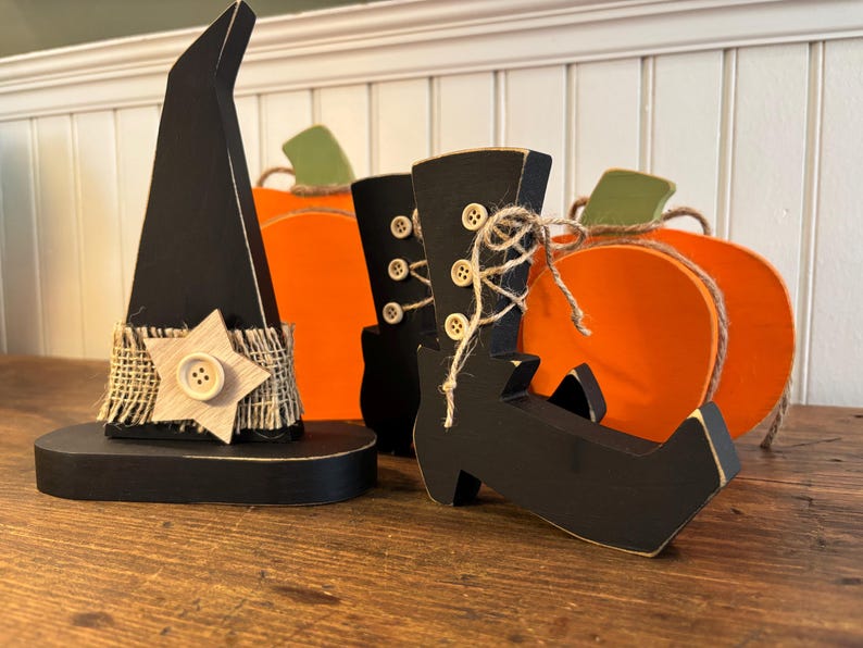 May include: Wooden Halloween decorations including a black witch hat with a star, black witch boots, and orange pumpkins. The hat has a burlap band with a wooden star and button. The boots have button details and twine. The pumpkins have green stems.
