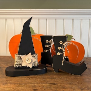May include: Wooden Halloween decorations featuring a black witch hat with a burlap and star accent, black boots with button details, and orange pumpkins. The hat and boots are on a wooden surface.