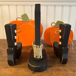 May include: A set of three black wooden boot-shaped decorations with white button accents. Two orange pumpkin cutouts with green stems and twine are in the background. The central boot has a burlap accent.