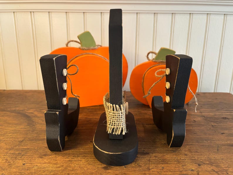 May include: A set of three black wooden boot-shaped decorations with white button accents. Two orange pumpkin cutouts with green stems and twine are in the background. The central boot has a burlap accent.