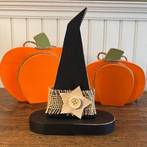 May include: A black wooden witch hat with a burlap band and a wooden star with a button. Two orange wooden pumpkins with green stems are in the background. Halloween decor.
