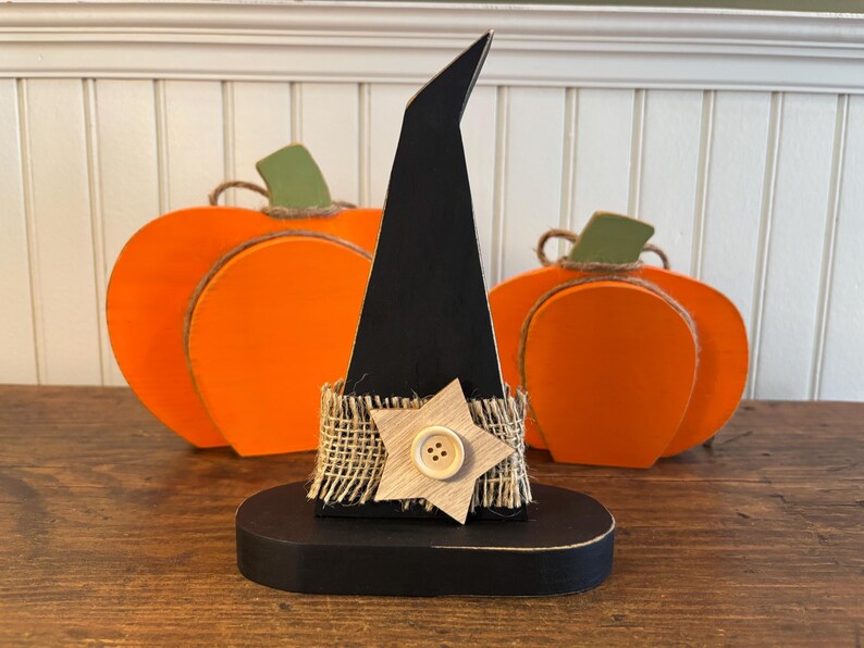 May include: A black wooden witch hat with a burlap band and a wooden star with a button. Two orange wooden pumpkins with green stems are in the background. Halloween decor.
