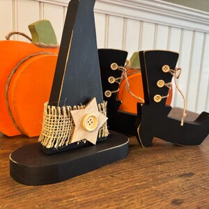 May include: Wooden Halloween decorations including a black witch hat with a star, a pair of black boots with button details, and an orange pumpkin. The hat has a burlap band and a wooden star.