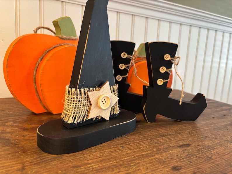 May include: Wooden Halloween decorations including a black witch hat with a star, a pair of black boots with button details, and an orange pumpkin. The hat has a burlap band and a wooden star.