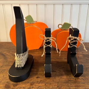 May include: Wooden Halloween decorations featuring a black witch hat, witch legs, and pumpkins. The witch hat has a burlap band, and the witch legs are tied with twine. The pumpkins are orange with green stems.
