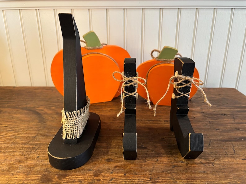 May include: Wooden Halloween decorations featuring a black witch hat, witch legs, and pumpkins. The witch hat has a burlap band, and the witch legs are tied with twine. The pumpkins are orange with green stems.