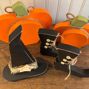 May include: Wooden Halloween decorations featuring two orange pumpkins with green stems, a black witch hat, and two black boots with button details. The hat has a decorative band, and the boots have twine accents.