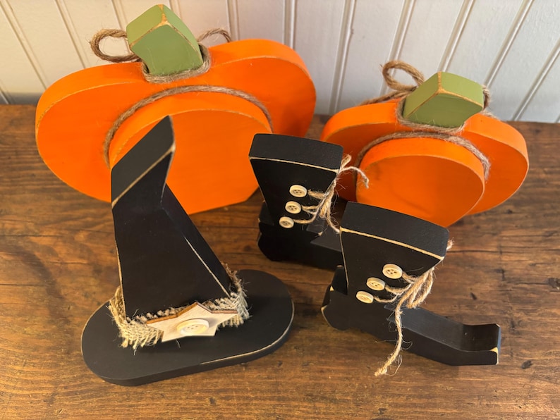 May include: Wooden Halloween decorations featuring two orange pumpkins with green stems, a black witch hat, and two black boots with button details. The hat has a decorative band, and the boots have twine accents.