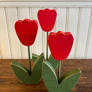 May include: Wooden tulip decoration with three red tulip heads with gold edging, green stems, and green leaves. The decorative flowers are arranged on a wooden surface, creating a charming spring or garden theme.