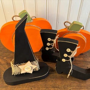 May include: Wooden Halloween decorations including an orange pumpkin, black witch hat with a burlap band and star, and black witch boots. The boots have button and twine details. The pumpkin has a green stem.