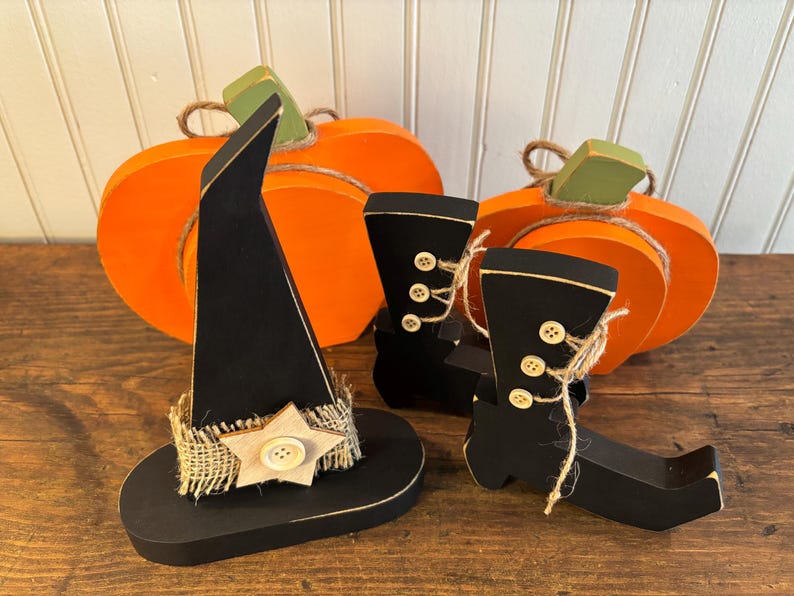May include: Wooden Halloween decorations including an orange pumpkin, black witch hat with a burlap band and star, and black witch boots. The boots have button and twine details. The pumpkin has a green stem.