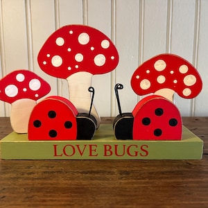 Pode incluir: Conjunto decorativo de madeira com duas joaninhas e três cogumelos. As joaninhas são vermelhas com pintas pretas, e os cogumelos têm chapéus vermelhos com pintas brancas. A base é verde com a inscrição "LOVE BUGS".