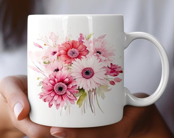 Könnte beinhalten: Weiße Keramiktasse mit einem Aquarell-Blumendesign. Das Design umfasst rosa, rote und weiße Gerberas und andere Blumen. Die Tasse hat einen gebogenen Henkel und wird von einer Person gehalten.