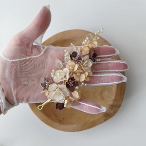Puede incluir: Un accesorio floral para el cabello con flores blancas, beige y moradas oscuras, acentuado con pequeñas perlas y alambre dorado. Un elegante adorno para bodas.